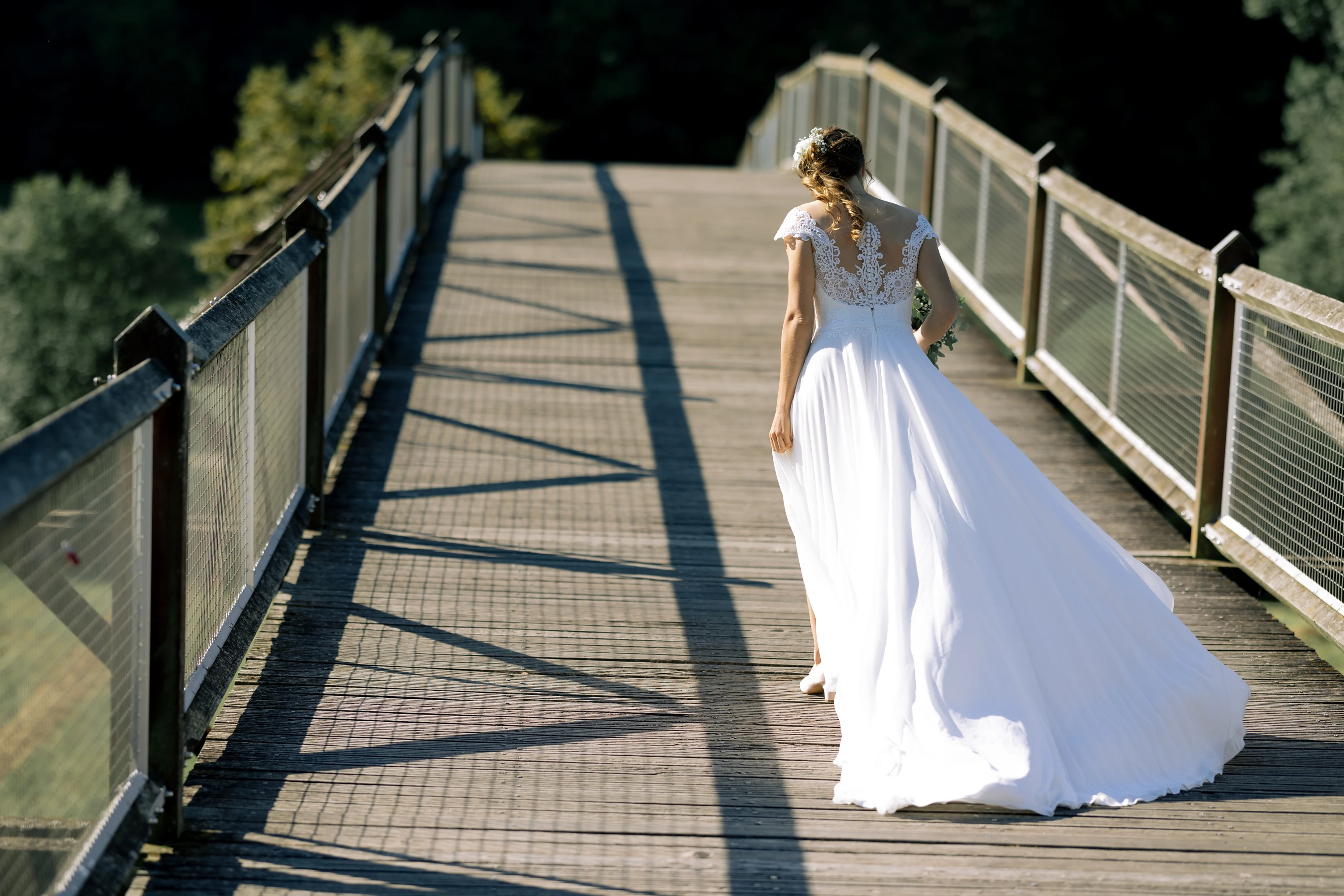 Die Braut geht auf einer Holzbrücke von der Kamera weg, das Kleid wellt sich im Wind.